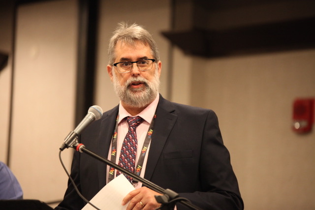 Man in suit presenting at a conference, standing at a microphone with a paper in hand, glasses and a beard.